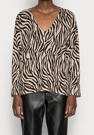 Personne portant un haut à manches longues col en V imprimé zèbre beige et noir et un pantalon en cuir noir, debout devant un fond uni.