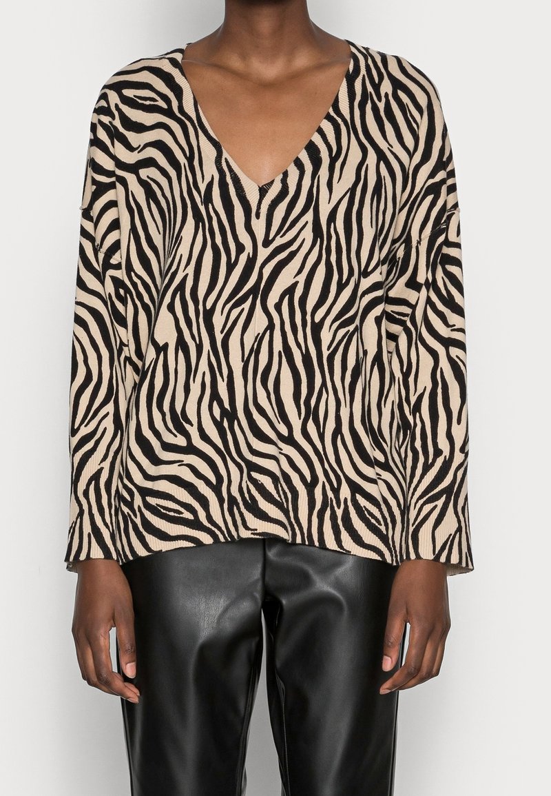 Personne portant un haut à manches longues col en V imprimé zèbre beige et noir et un pantalon en cuir noir, debout devant un fond uni.
