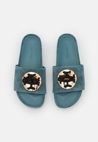 Paire de sandales slides en daim bleu sarcelle avec de grands emblèmes décoratifs dorés et noirs sur les brides, vues de dessus sur fond blanc.