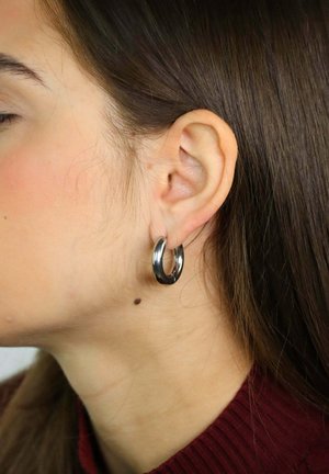 Zijaanzicht van een persoon met lang bruin haar die een zilveren ringoorbel en een donkerrood geribbeld topje draagt.