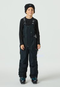 Svarta skidbyxor med hängslen och justerbara remmar, ficka med dragkedja fram och dragkedjor på sidorna. Bärs över en svart långärmad tröja och kombineras med blå skidpjäxor.