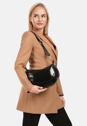 Femme blonde en blazer camel tenant un sac à main en cuir noir avec des clous métalliques sur l'épaule, sur fond blanc.