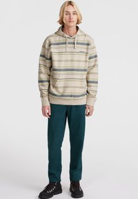 Sudadera de rayas beige con un bolsillo delantero, que presenta líneas horizontales azules y verdes. Combinada con pantalones verde oscuro y botas negras.