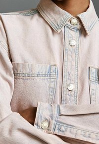 Veste en denim rose clair avec un col classique, des boutons-pression argentés et deux poches poitrine. Présente des coutures contrastantes et une texture douce.
