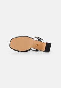 Zwarte haksandaal met brede band en enkelgesp. Getextureerde beige zool met een kleine zwarte hak. Gouden logo-detail nabij de hak.