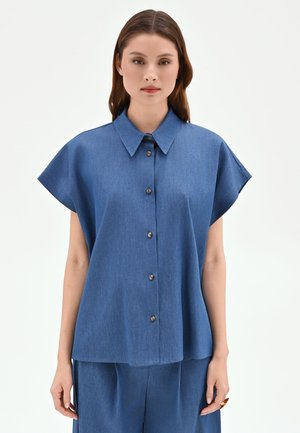 Femme portant une chemise bleue ample à boutons avec manches courtes et pantalon bleu assorti, debout devant un fond uni.