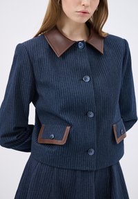 Námořnický pruhovaný saka s hnědým koženým límcem, se dvěma předními kapsami a modrými knoflíky. Strukturovaný design s viditelným švem.