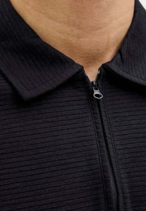 Giacca nera con zip centrale, caratterizzata da una texture a coste e un colletto ripiegato. La zip è funzionale nel design.
