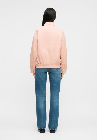 Γυναίκα με μακριά μαύρα μαλλιά, φορόντας ένα ανοιχτό ροζ μπουφάν και μπλε τζιν, γυρισμένη με την πλάτη προς το απλό ανοιχτό φόντο.