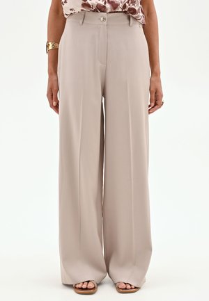 Pantalon beige à jambes larges avec bouton et fermeture éclair devant, porté avec un haut fleuri, un bracelet en or, une bague et des sandales ouvertes.