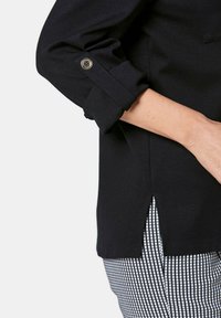 Schwarzes Hemd mit hochgeschlagenen Ärmeln und einem Knopfdetail. Seitlicher Schlitzsaum. Getragen über gemusterten schwarz-weißen karierten Hosen.