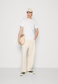Hvit bomulls t-skjorte, beige vide bukser, svart-hvite joggesko, og en beige caps. T-skjorten har en liten logo på brystet.
