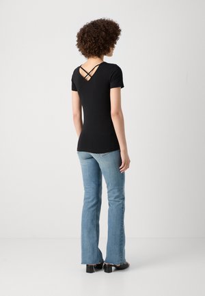 Kvinde med krøllet hår iført en sort kortærmet top med krydsede stropper på ryggen og lyseblå, vidde jeans stående på en hvid baggrund.