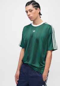 Mladić s pletenicama, odjeven u zelenu Adidas majicu s bijelim prugama i tamnoplave Adidas hlače, stoji s jednom rukom u džepu.