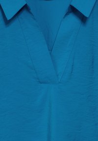 Türoises Bluse mit V-Ausschnitt, kurzem Kragen und gerauter Textur, die sichtbare Nähte entlang der Nähte aufweist.