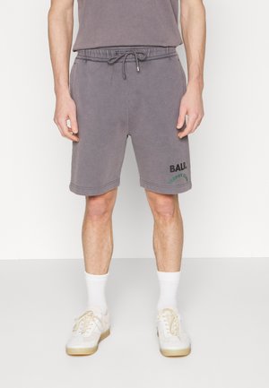 Mand iført grå snørelommer med "BALR." og grøn tekst på venstre ben, kombineret med hvide sokker og off-white sneakers.