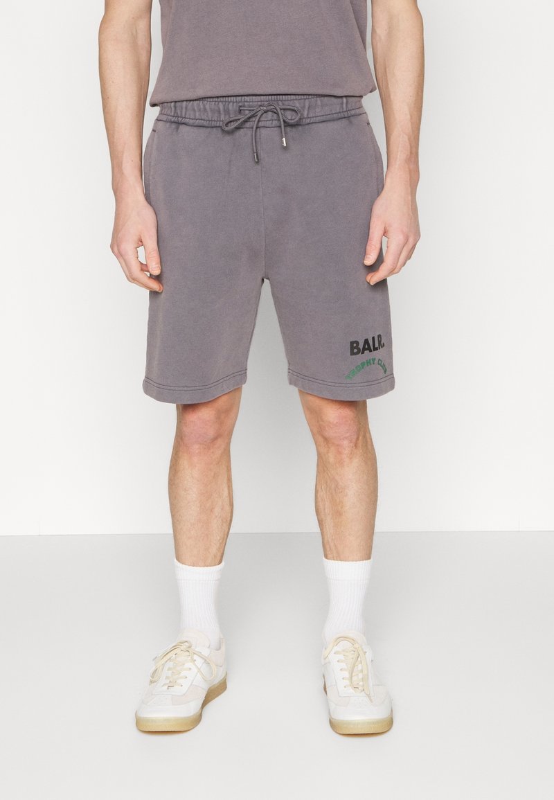 Homme portant un short gris à cordon avec l'inscription "BALR." et du texte vert sur la jambe gauche, assorti à des chaussettes blanches et des baskets crème.