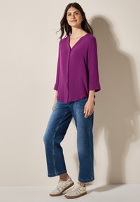 Blouse violette à boutons, avec un col en V et des manches trois-quarts, associée à un jean large bleu clair et des baskets beiges avec des accents blancs.