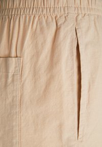 Beiga linnebyxor med elastisk midja, med en sidficka och avslappnad passform. Texturen verkar lätt och något rynkad.