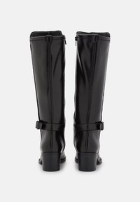Bottes noires en cuir montantes jusqu'au genou avec une finition lisse, dotées d'une fermeture éclair et de brides réglables à la cheville, sur un talon empilé.