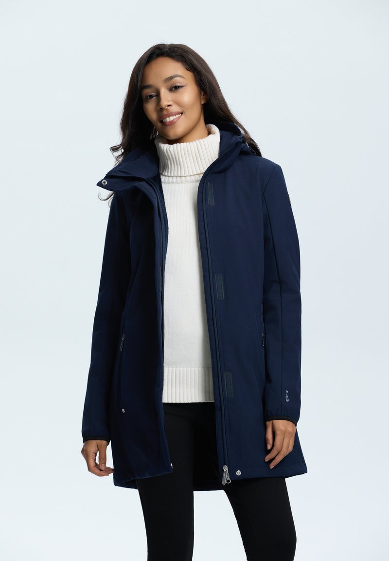 Cappotto lungo blu navy con cappuccio, dotato di chiusura con zip, due tasche laterali e un morbido strato interno. Indossato sopra un maglione a collo alto bianco.