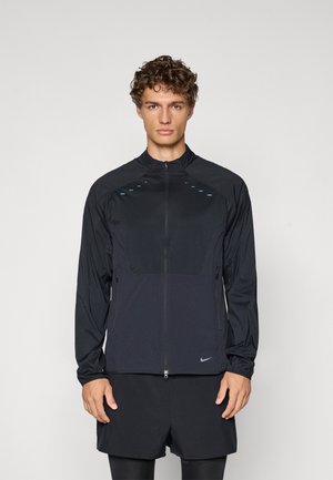 Svart, lett, langermet treningjakke med glidelås foran, to sidelommer, blå reflekterende detaljer og Nike-logo på kanten.