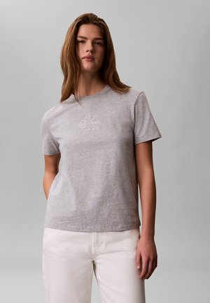 Νεαρό άτομο με μακριά μαλλιά που φοράει ένα ανοιχτό γκρι t-shirt Calvin Klein και λευκό παντελόνι, στέκεται μπροστά σε μονόχρωμο γκρι φόντο.