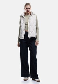 Polstret, lysebeige vest med lynlås foran og hætte. Kombineret med en tætsiddende grøn top og brede mørke jeans. Modellen står på en hvid baggrund.