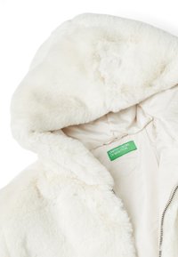 Giacca in pelliccia sintetica bianca con cappuccio, caratterizzata da una texture morbida, chiusura con zip e un'etichetta del marchio verde sull'imbottitura interna.