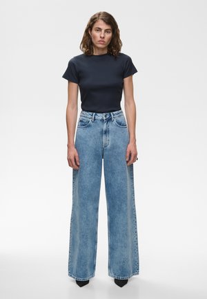 Femme aux cheveux bruns mi-longs portant un haut noir à manches courtes rentré dans un jean large taille haute bleu clair avec des chaussures noires.