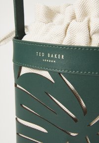 Grüne Tasche aus Kunstleder mit ausgeschnittenem Blättermuster, ausgestattet mit einer beigen, strukturierten Zugbandschicht und goldener Logodetaillierung.
