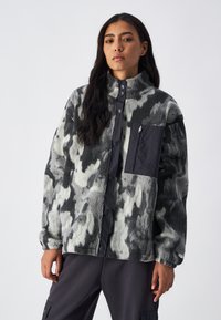Giacca in pile oversize e morbido, con motivo mimetico bianco e nero, dotata di tasca sul petto con zip e chiusura a bottoni a pressione.