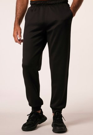 Zwarte sweatpants van een gladde stof, met een elastische tailleband, taps toelopende boorden en zijzakken. Draagt met zwarte sneakers.