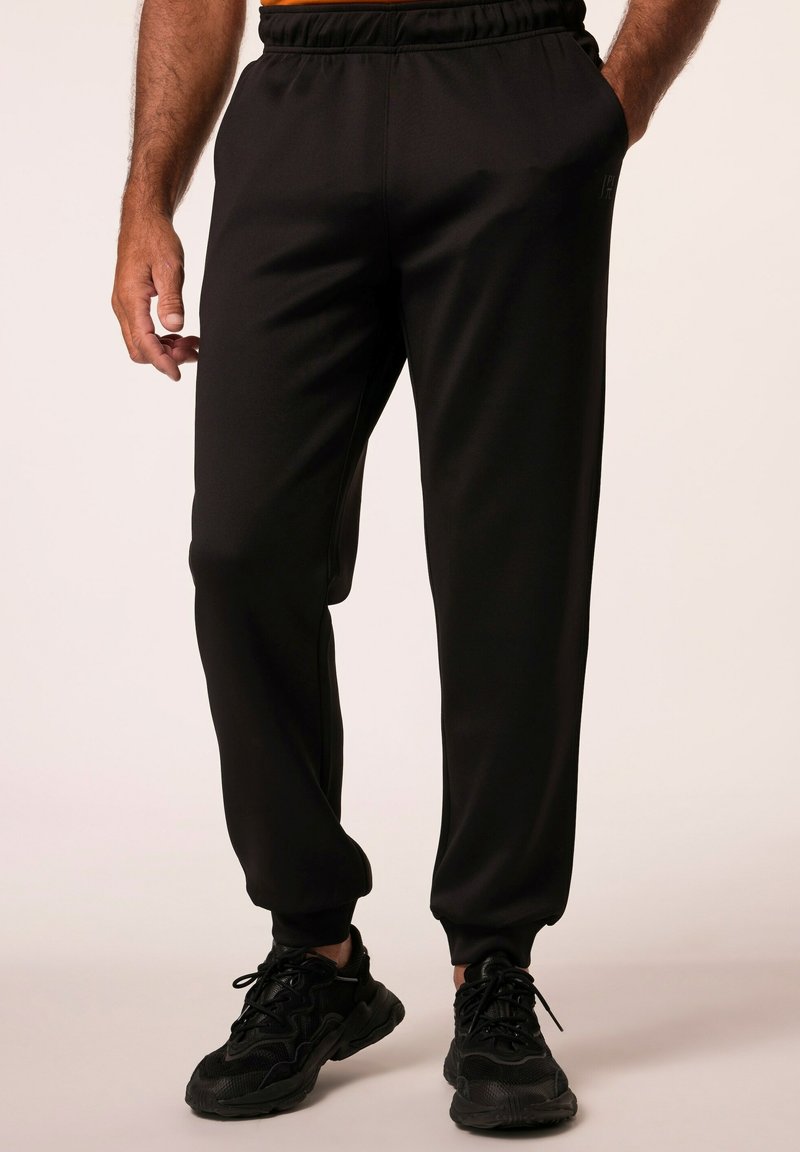 Pantalones sudorosos negros fabricados en tela suave, con una cintura elástica, puños ajustados y bolsillos laterales. Llevados con zapatillas negras.