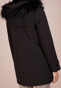 Svart parkas med avtagbar fuskpälshood, med rak skärning, långa ärmar, knäppta manschetter och utan synliga mönster eller detaljer.