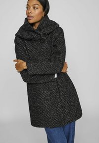 Manteau noir texturé avec un grand col et une fermeture à bouton. Coupe ample avec des manches longues et une finition droite, superposé sur un jean bleu.
