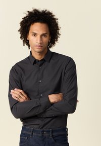 Camicia nera con bottoni, dal tessuto liscio, maniche lunghe e leggera lucentezza. Presenta un colletto classico e chiusura frontale con bottoni. Sono visibili anche jeans in denim scuri.