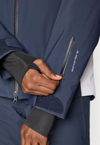 Tengerészeti vízálló dzseki, cipzáros zsebbel a hüvelyen, tépőzáras mandzsettával és rugalmas belső ujjal. Az ujjon logóval ellátva.