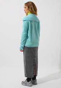 Helles bläuliches Oversized-Pullover mit breitem Kragen, kombiniert mit einem grauen Midirock mit Schlitz hinten und grauen Sportschuhen.