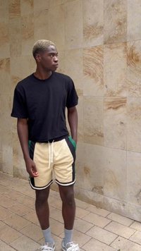 Svart kortermet t-skjorte, kremfargede shorts med grønne detaljer og svart kant, grå sokker og joggesko. Står mot en teksturert vegg.