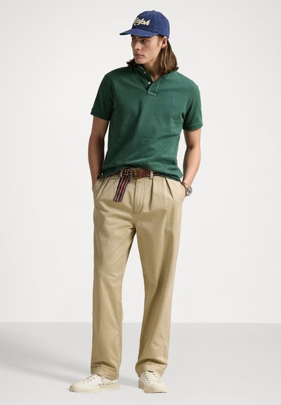 Groene polo met een twee-knopsluiting, beige broek en een blauwe pet. De pet heeft borduursel; de riem heeft meerkleurige strepen.