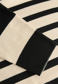 Schwarz- und cremefarbener gestreifter Pullover, der eine gerippte Textur an den Bündchen und am Körper zeigt, mit abwechselnd breiten und schmalen Streifen.