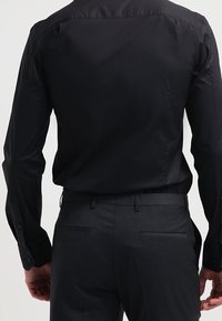 Uomo che indossa una camicia nera a maniche lunghe infilata in pantaloni eleganti grigio scuro, visto da dietro su uno sfondo neutro.