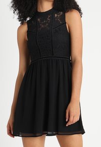 Robe noire avec un corsage en dentelle, un col haut et un design sans manches. La jupe fluide présente un ourlet légèrement transparent. Texture lisse tout au long.
