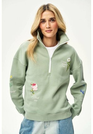 Blondynka w zielonym sweatshirt z półzapinanym suwakiem, ozdobionym kwiatową haftem i napisem, w połączeniu z jasnoniebieskimi dżinsami, pozująca z rękami w kieszeniach.