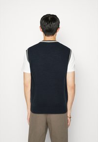 Granatowa dzianinowa kamizelka z prążkowanym dołem i białym akcentem przy dekolcie, noszona na białej koszuli. Tył jest gładki i teksturowany.