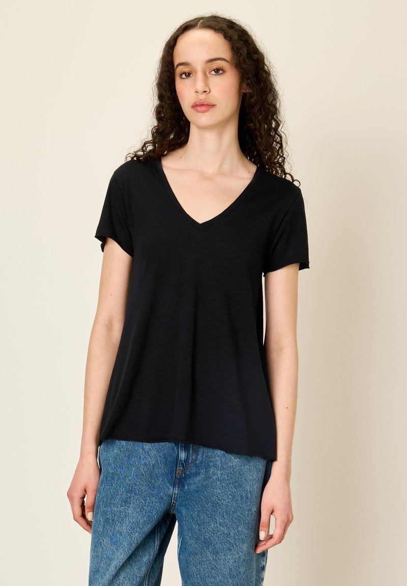 Woman with curly hair wearing a black V-neck short-sleeve shirt and blue jeans, standing against a beige background.