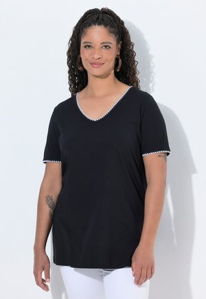 Frau mit lockigem Haar, die ein schwarzes V-Ausschnitt-Shirt mit weißem Rand und weiße Hose trägt, steht vor einem schlichten hellen Hintergrund.