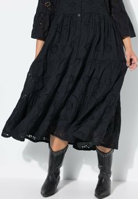Zwarte katoenen maxi-jurk met geborduurde bloemen uitsparingen, knoopsluiting aan de voorkant en een gelaagd ontwerp. Gekoppeld aan zwarte puntige laarzen met studaccenten.