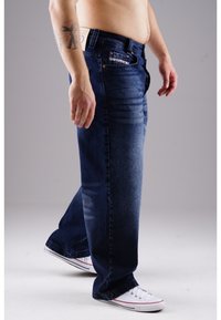 Dunkelblaue Jeans mit geradem Schnitt, die ein ausgewaschenes Muster aufweisen. Die Hose hat sichtbare Nähte und ein Fünf-Taschen-Design und wird mit weißen Sneakern kombiniert.